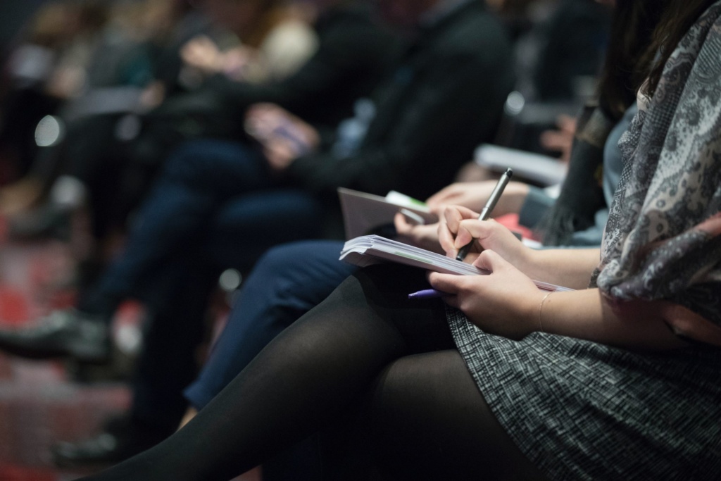 Person writing notes A side-angle, close-up shot of a person's hands holding a pen and notebook, actively writing during a conference, lecture, or meeting, with a blurred audience in the background.