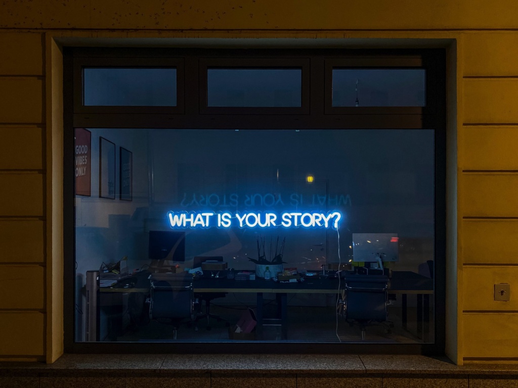 Neon story sign A bright blue neon sign inside a dark office window reads, "WHAT IS YOUR STORY?" with a faint reflection of the question visible on the glass.
