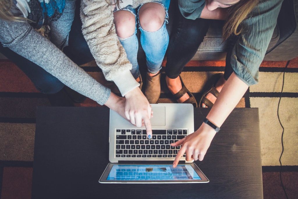 Three people, seen from above, lean over a laptop and a tablet. Two hands point at the screens, one at the keyboard, and another at the tablet, suggesting collaboration or group work.