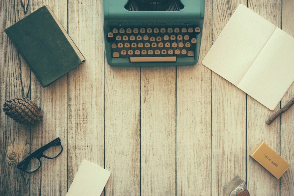 An overhead shot of a light-colored wooden desk with a teal vintage typewriter at the top center. Surrounding the typewriter are various items, including a closed book, an open notebook, a pinecone, a pair of black glasses, and a pencil.