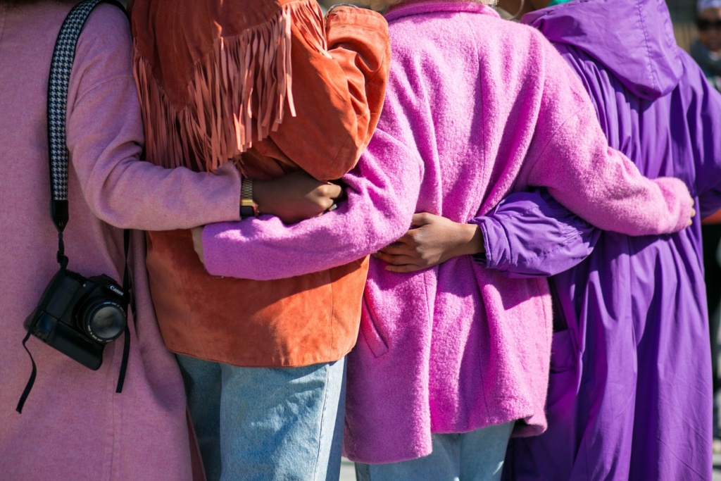 Group of people in colorful coats A group of people stand with their arms wrapped around each other's shoulders and waists. They are wearing colorful, textured coats in shades of pink, purple, and orange.