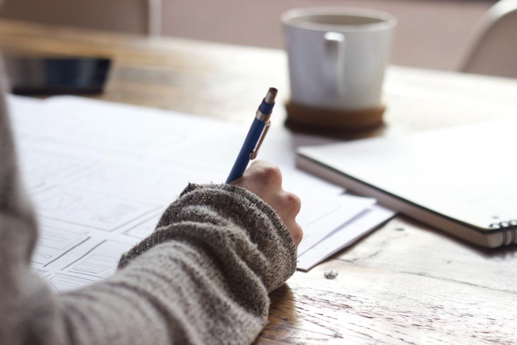 Writing notes to develop a creative brief. A person writing notes on paper with a pen at a desk, next to a coffee cup and notebook.