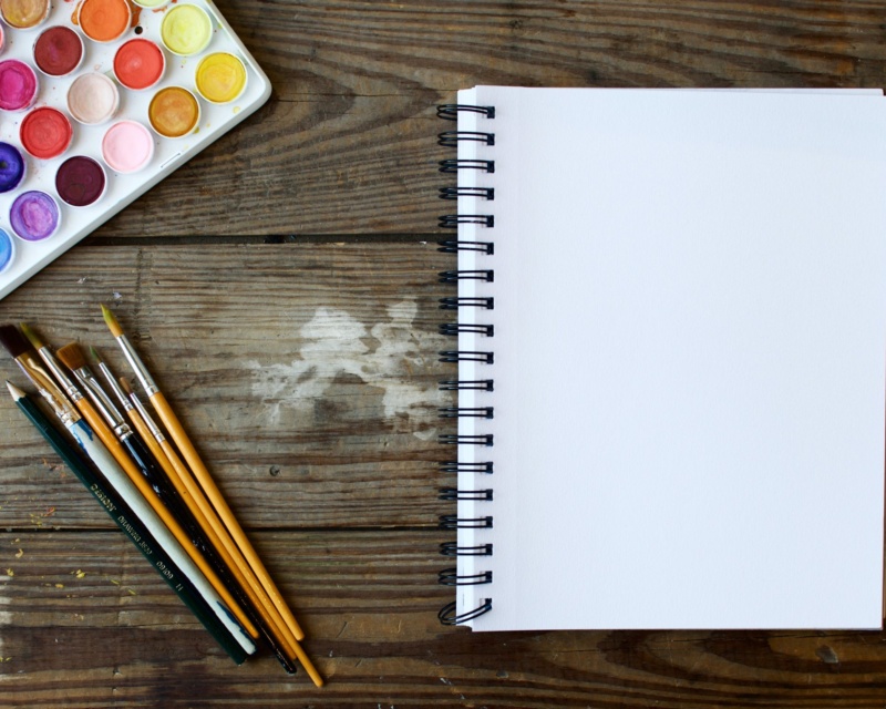 Blank page of spiral notebook next to pens, pencils and a tray of watercolors symbolizing starting of a creative brief