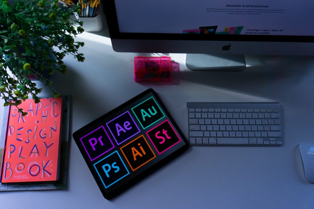 Graphic design workspace with Adobe Creative Suite tools Workspace with a tablet displaying Adobe Creative Suite icons, a Graphic Design Playbook, and an Apple computer setup.
