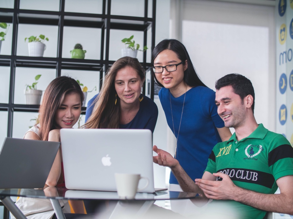 A group of four colleagues gathered around a laptop, smiling and discussing work together.