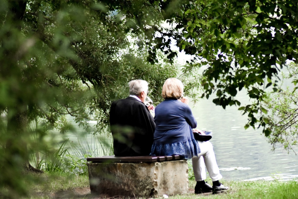 Senior couple sitting on a bench overlooking a lake while each is having a glass of wine.