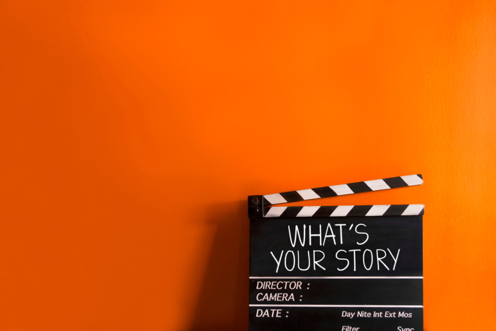 What’s Your Story clapperboard on orange background Black film clapperboard with the words "What’s Your Story" against a bright orange background.