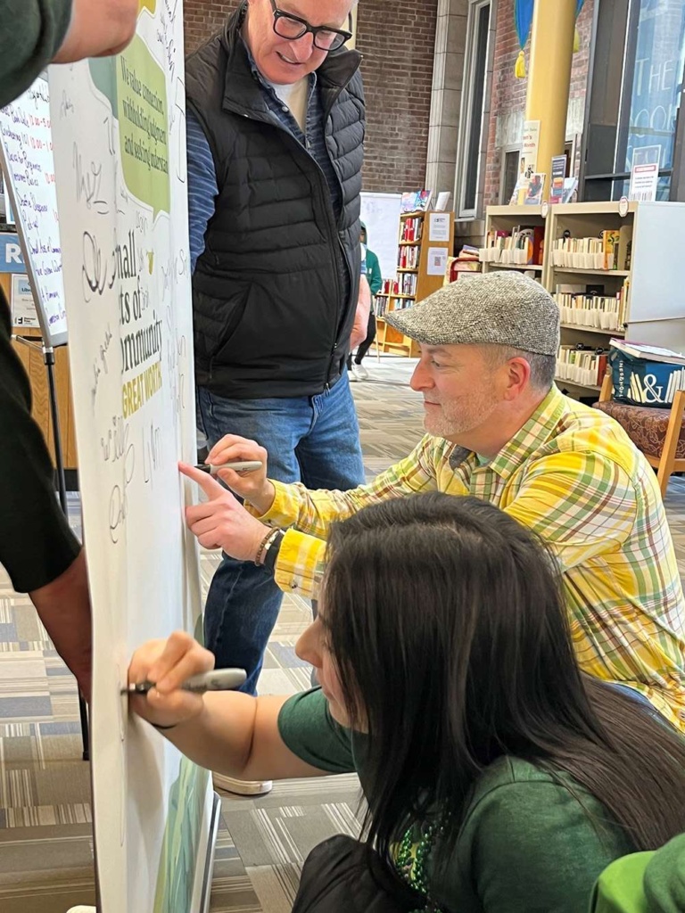 Mark Crow, President of Tenth Crow Creative, stands beside community members signing a campaign board during the Small Acts of Community initiative.