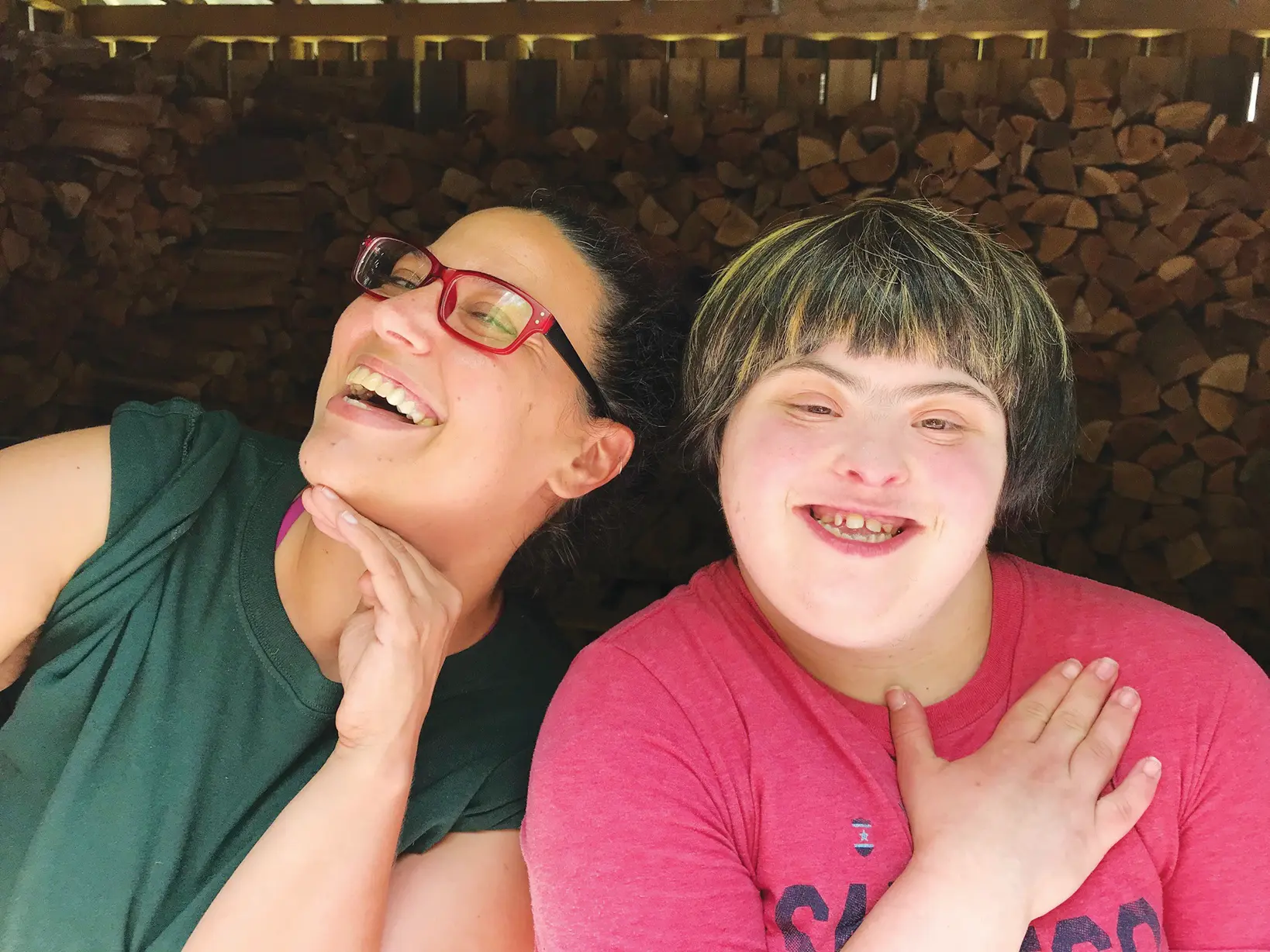 Two people smiling at the camera in front of chopped wood. One in the intellectual and developmental disabilities program.